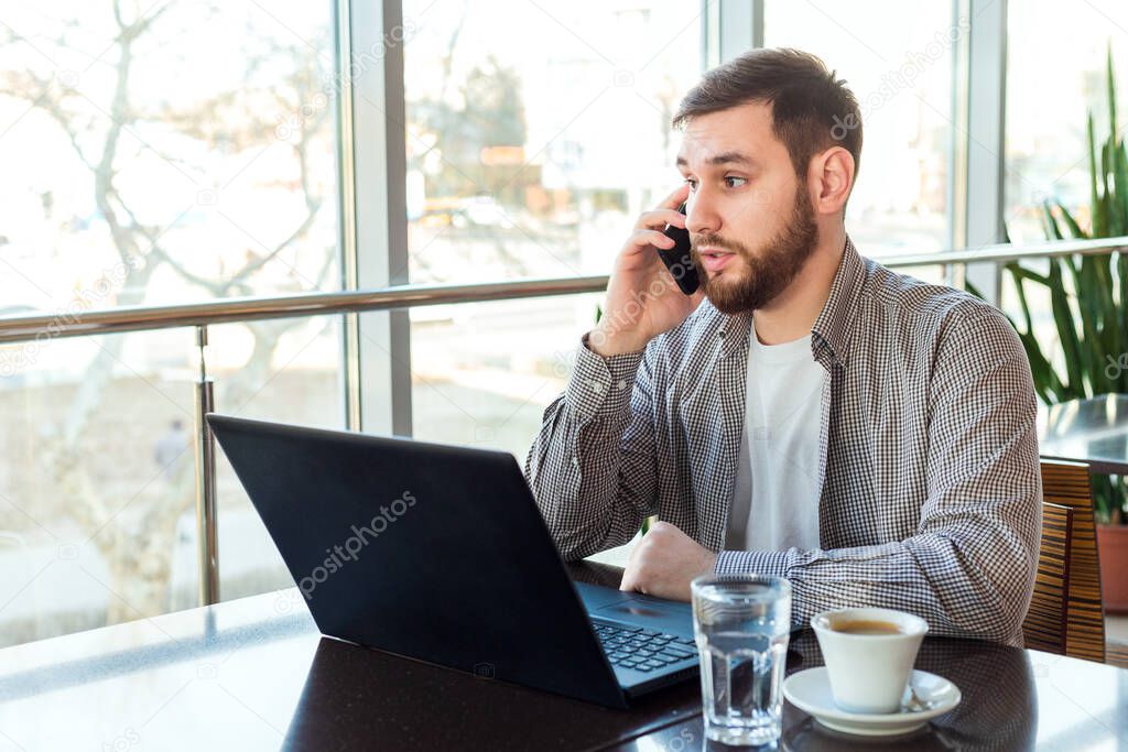 Phone conversation. Upset young man talking over phone disappointed hear bad news. Portrait tired, exhausted man in cafe or modern office with smartphone and laptop. Attractive businessman freelancer.