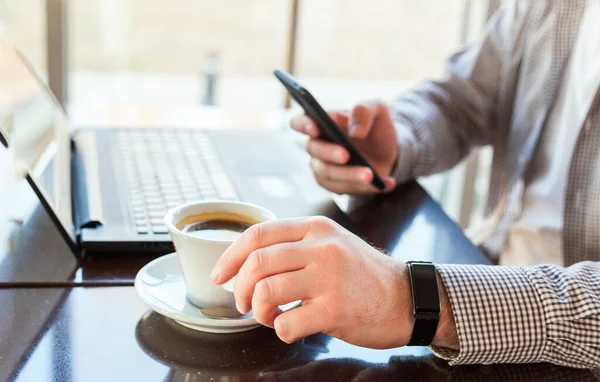 Man in smart watch drinking coffee on work space. Man using laptop, holding smart phone for business work or study.Close up smart watch on male hand. Freelancer works in cafe, office, texting massage.