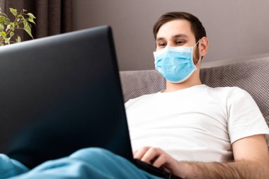 Ev ofisinden çalışan genç beyaz bir adam bilgisayar ve internet kullanarak koruyucu maske takıyor. Coronavirus covid 19 karantinası. Uzaktan çalışma, serbest çalışma, kanepede rahat ofis çalışma yeri..