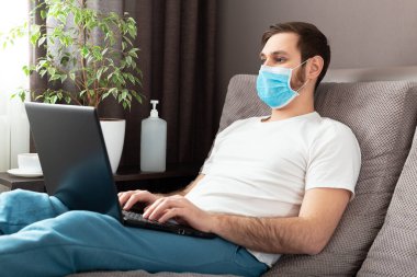 Genç beyaz adam evde çalışıyor koruyucu maske takıyor, dizüstü bilgisayar ve internet kullanıyor. Konforlu ofis, Coronavirus salgını sırasında kanepenin üzerinde çalışma yeri, karantina alanı 19. Uzak çalışma, serbest çalışan..