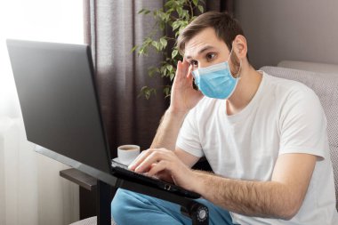 Ev ofisinden, dizüstü bilgisayar ve internet kullanarak koruyucu maske takan stresli, beyaz, genç bir adam. Coronavirus covid 19 karantina. Uzaktan çalışma, serbest çalışma, ev işyeri kanepesi..