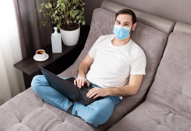 Mutlu, rahat, beyaz bir adam evde çalışıyor koruyucu maske takıyor ve dizüstü bilgisayar kullanıyor. Konforlu ofis, Coronavirus salgını sırasında kanepenin üzerinde çalışma yeri, karantina alanı 19. Uzak çalışma, serbest çalışan..
