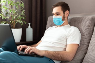 Genç beyaz adam evde çalışıyor koruyucu maske takıyor, dizüstü bilgisayar ve internet kullanıyor. Konforlu ofis, Coronavirus salgını sırasında kanepenin üzerinde çalışma yeri, karantina alanı 19. Uzak çalışma, serbest çalışan..