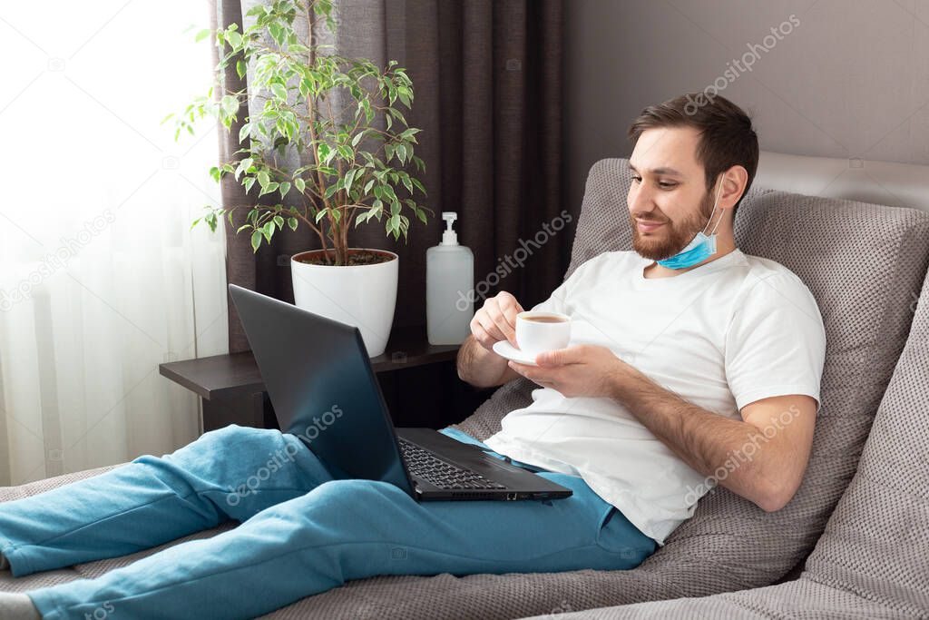 Happy young caucasian man in medical face mask drink tea while working from home using laptop. Cozy home office, workplace on sofa on coronavirus pandemic covid 19 quarantine. Remote work, freelancer.