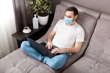 Ev işi yeri. Yüz cerrahisi maskeli adam evde dizüstü bilgisayar kullanarak çalışıyor. Kanepede rahat bir ofis. Coronavirus covid-19 sosyal mesafeli kişisel karantina, evde yalnız kalma kavramı.