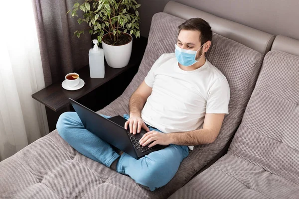 Home work place. Man freelancer in face surgical mask working from home using laptop. Cozy home office on sofa. Coronavirus covid-19 social distancing self quarantine, Stay home isolation concept.