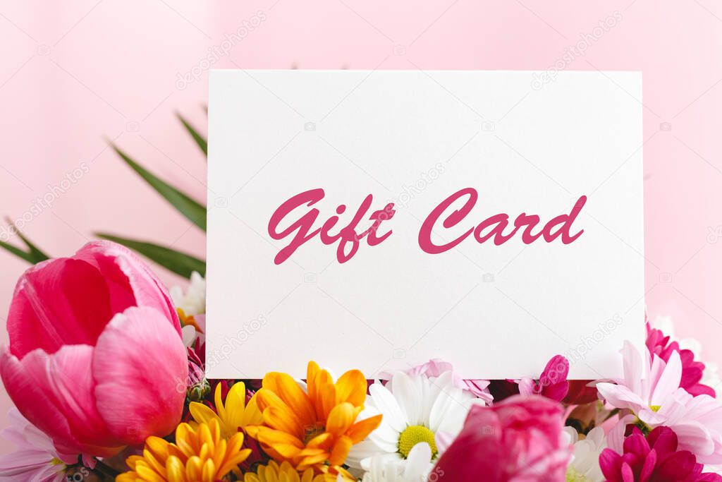 Gift card in flowers bouquet on pink background. Gift card present coupon for woman. Surprise voucher for Mother's Day, Happy Birthday or anniversary for mother day, daughter or granny.