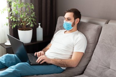 Genç beyaz adam evde çalışıyor koruyucu maske takıyor, dizüstü bilgisayar ve internet kullanıyor. Konforlu ofis, Coronavirus salgını sırasında kanepenin üzerinde çalışma yeri, karantina alanı 19. Uzak çalışma, serbest çalışan..