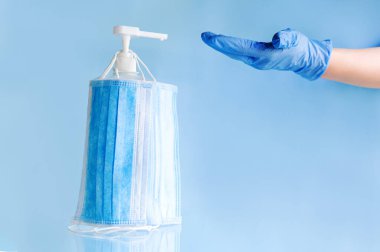 Tıbbi yüz maskeleri, eldivendeki doktor ellerini dezenfekte etmek için şişe alkolüne takılmış tek kullanımlık cerrahi maske. Coronavirus covid 19 virüs önleme, mavi arka planda el hijyeni kavramı.