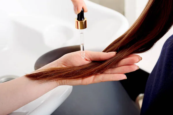Kadın eli, uzun, kahverengi saçlara yağ sürüyor. Saç bakım kozmetik ürünleri, banyo güzellik spa ürünleri. Tedavi için kadın saç serumu. Kuaför salonu, banyo gibi yerlere karşı saç uçları..