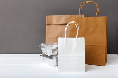 Beyaz el işi kağıt torbaları ve beyaz masa gri arka planında yiyecek kapları. Yemek dağıtım servisi. Folyo kutularındaki yiyecek paketleri, boş karton kağıt paketleri. Boşluğu kopyala.