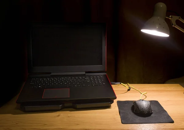 Laptop with a computer mouse on the table under the light of a desk lamp. Workplace at home and at work. Black laptop with red design and a wired mouse with the mat under the lamp.