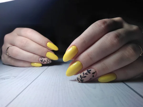 Yellow nail polish for women the handles with the design of the black twigs on camouflage coating. Beautiful manicure with matte yellow coating on long nails. Manicure work on a wooden table.