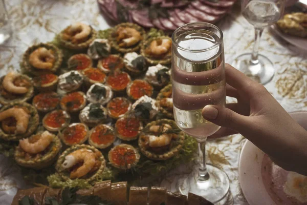 A woman holds a glass of sparkling champagne against the background of a festive table. A glass of sparkling wine in the girl\'s hand on the background of salads and tartlets with shrimp.