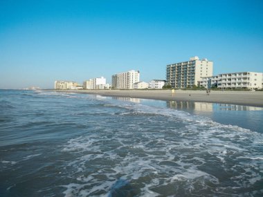 Tatil otelleri ve tatilcileri olan okyanus kıyısında. Beyaz kumlu deniz kıyısı, açık gökyüzü ve mavi deniz. Küçük dalgalar ve okyanusta sakinlik. Tatilciler sahil boyunca yürür..