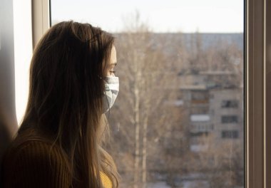 Maskeli bir kız pencere pervazına oturur ve pencereden şehre bakar. Tıbbi maskeli bir kadın karantinada oturuyor. Karantina ve kendini izole etme kavramı..