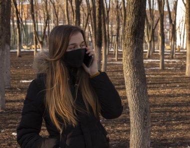Siyah koruyucu maskeli bir kız parkta yürüyor. Tıbbi maskeli bir kadın telefonda konuşuyor. Coronavirüs salgını. Covid-19 'dan koruma.