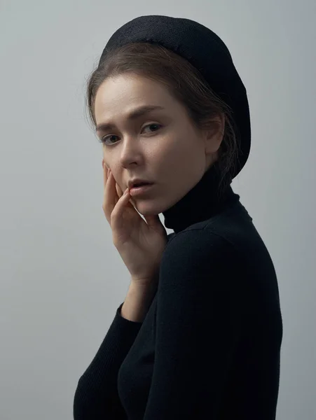 Drammatico ritratto di una giovane bella ragazza con lentiggini in un dolcevita nero e un cappello sulla testa su uno sfondo bianco in studio — Foto Stock