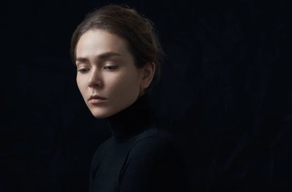 Dramatic portrait of a young beautiful girl with freckles in a black turtleneck on black background in studio