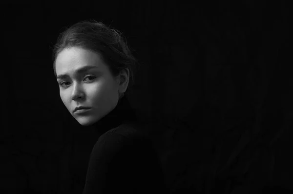 Dramatic black and white portrait of young beautiful girl with freckles in a black turtleneck on black background in studio