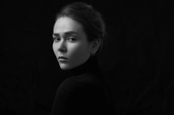 Dramatic black and white portrait of young beautiful girl with freckles in a black turtleneck on black background in studio