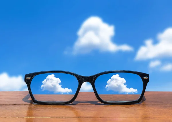 Gafas graduadas sobre mesa de madera frente a nubes blancas sobre fondo azul cielo — Foto de Stock