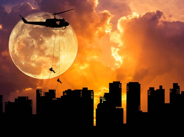 Silhouette soldiers in action rappelling climb down from helicopter on building skyscraper on sunset with full moon — Stock Photo, Image