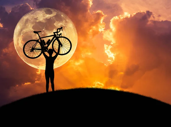 Silhueta o homem levantando bicicleta acima de sua cabeça no pôr do sol com fundo de lua cheia . — Fotografia de Stock