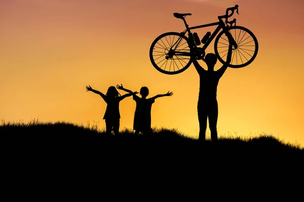 Silhueta o homem levantar bicicleta acima de sua cabeça no prado com as crianças levantar as mãos para cima no pôr do sol . — Fotografia de Stock