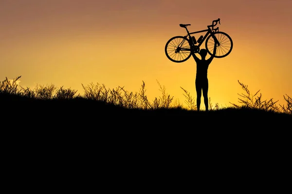 Silhueta o homem ficar em ação levantando bicicleta acima de sua cabeça com o pôr do sol — Fotografia de Stock