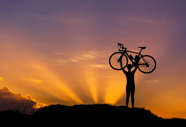 Silhueta o homem ficar em ação levantando bicicleta acima de sua cabeça ao pôr do sol — Fotografia de Stock