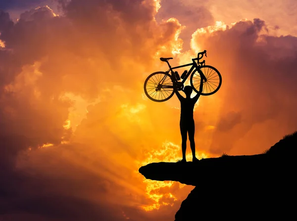 Silhueta o homem em ação levantando bicicleta acima de sua cabeça no topo da montanha com o pôr do sol — Fotografia de Stock