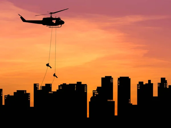 Soldados silhueta em ação rapel descer de helicóptero na construção de arranha-céus ao pôr-do-sol — Fotografia de Stock