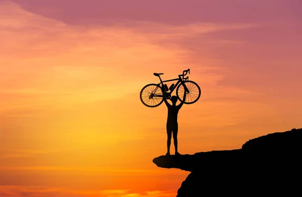 Silhueta o homem que levanta a bicicleta acima de sua cabeça no alto da montanha do penhasco da rocha com sunse — Fotografia de Stock