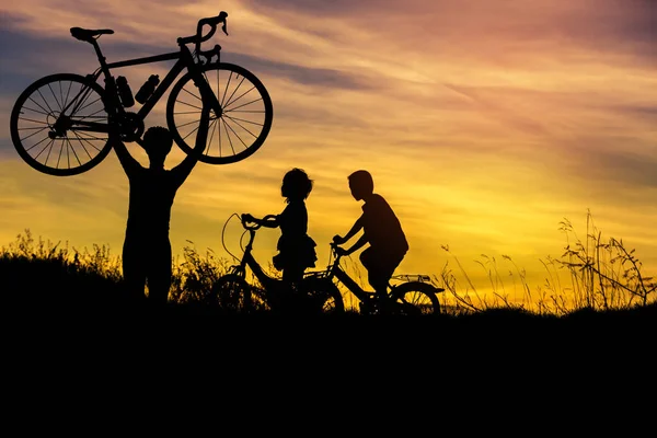 Silhueta o homem levantando bicicleta acima de sua cabeça com menino e menina se divertindo andando de bicicleta no pôr do sol — Fotografia de Stock