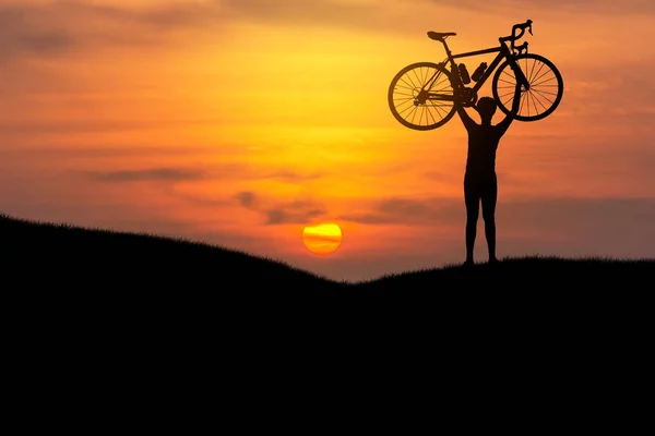 Silhueta o homem ficar em ação levantando bicicleta acima de sua cabeça ao pôr do sol — Fotografia de Stock