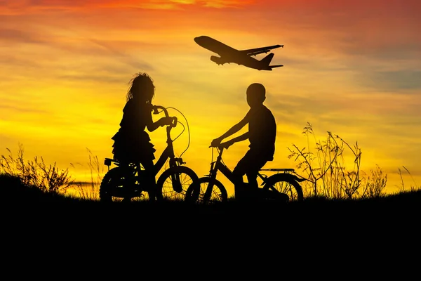 Силует маленький хлопчик і маленька дівчинка їде на велосипеді дивитися на літак, що летить на заході сонця — стокове фото