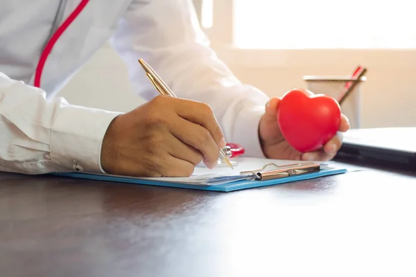 Doctor writing prescription on paper and holding red heart on wo