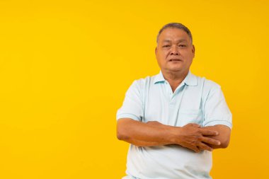 Taylandlı özgüvenli yaşlı adam kollarını kavuşturup stüdyodaki sarı arka plandaki kameraya bakıyor.