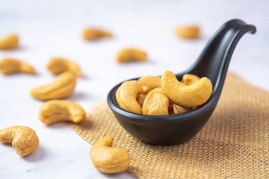 Kavrulmuş kaju fıstığı siyah kaşıkta tuzlu beyaz masada.