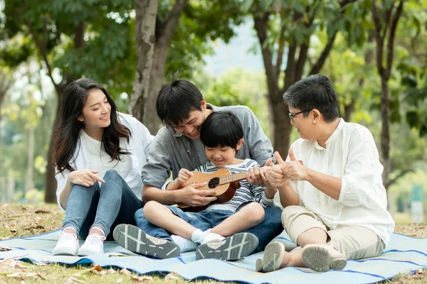 Happy family with grandma, mom with dad teaching son  playing guitar and sing a song in park, Enjoy and relax people picnic outside