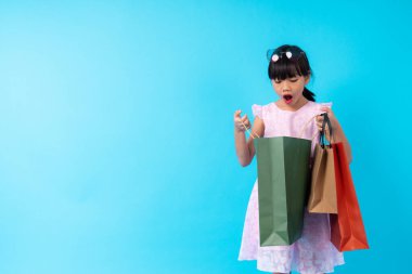 Mutlu Asyalı kız çocuğu şık bir şekilde alışveriş çantasına bakıyor ve vay canına, stüdyoda mavi arka plana şaşırmış, çocuk modası konsepti için para ödenen bir yaşam tarzı.