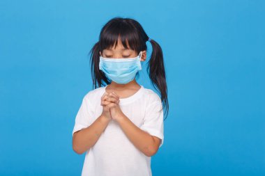 PM2.5 ve Coronavirus Covid-19 'u korumak için maske takan Asyalı bir kız. Pazar günü GodD.Online kilisesine dua etmek için evde kal. Küçük Asyalı kız evde dua ediyor. Sosyal uzaklık. Mavi arka planda.