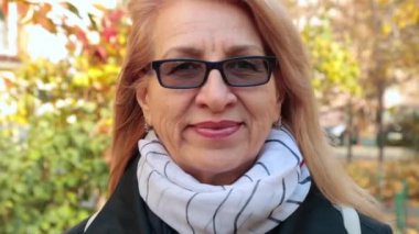 Gülümseyen gözlüklü neşeli olgun kadın. Trençkotlu, uzun saçlı, gülümseyen yetişkin bir kadın. Açık havada. Yaşlılar ve iletişim kavramı. yakın plan