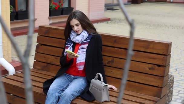 一个快乐的女人坐在长椅上 用手机发短信 美丽的黑发女人在户外使用智能手机 她又笑又笑 — 图库视频影像