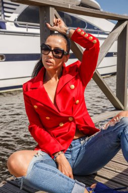 Güzel genç kadın güneş gözlüğü ve arka plan üzerinde yat ile ahşap iskele üzerinde oturan kırmızı ceket