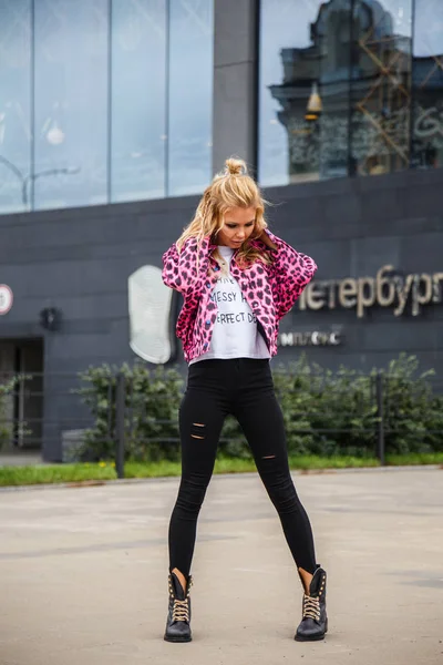 Pembe ceket ve açık havada ile ayakta siyah kot giyen güzel sarışın kadın alışveriş arka plan üzerinde bulanık