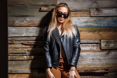 Genç sarışın kız stüdyo çekim. Güzellik kadını şık sütyen, ceket ve grunge ahşap duvar arka plan üzerinde duran güneş gözlüğü 