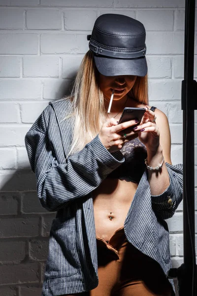 ブラとタバコを喫煙とスマート フォン 白いレンガ壁背景に Selfie を作るスタイリッシュなキャップでブロンドの女の子のスタジオ ポートレート — ストック写真