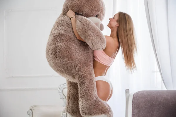 スポーティなボディを持つ美しい女の子のスタジオ撮影 若いセクシーな女性ランジェリーに立って 大きなクマのぬいぐるみを抱いてします ホーム インテリアの背景 — ストック写真